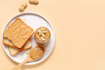 Jar with tasty peanut butter, wooden spoon, toasts and nuts on beige background