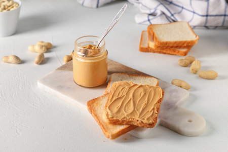 Jar of tasty peanut butter with spoon, toasts and nuts on white background