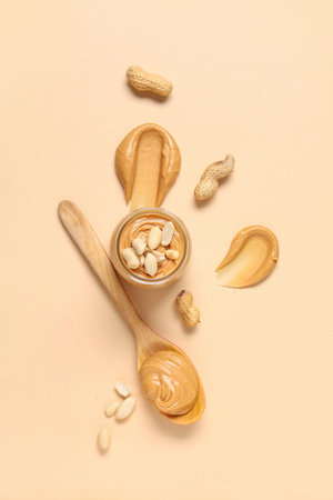 Jar with tasty peanut butter, wooden spoon and nuts on beige background