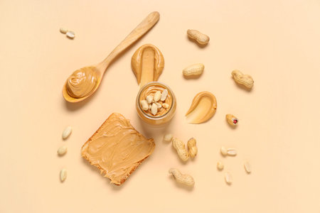 Jar with tasty peanut butter, wooden spoon, toast and nuts on beige background