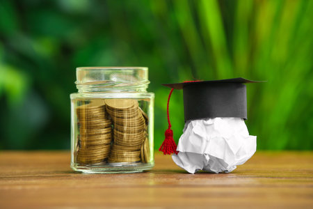 Crumpled paper with graduation hat and jar of coins on table outdoors. Student loan conceptの写真素材