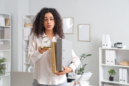 Stressed African-American businesswoman with alarm clock and folders in office. Time management...