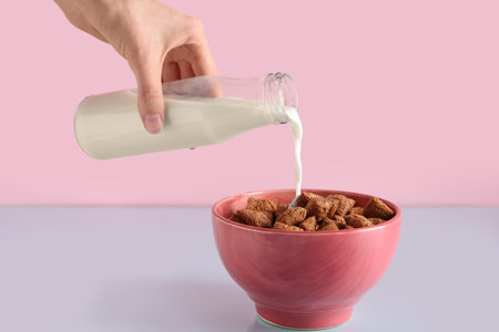 Young woman pouring milk into bowl with healthy cornflakes on color background