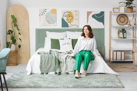 Young woman sitting near stylish paintings on light wall in bedroom