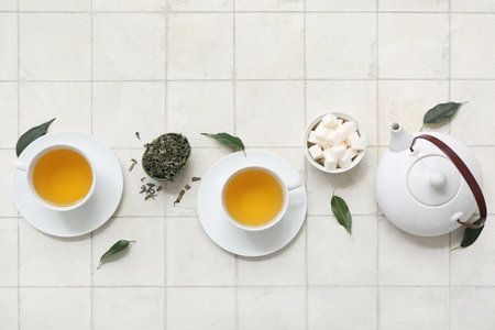 Cups of hot green tea with sugar cubes and dried leaves on white tile backgroundの写真素材