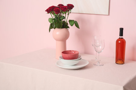 Vase with roses, dishes and wine on tablecloth near pink wall in roomの写真素材