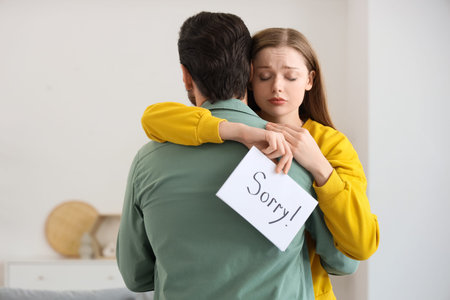 Sad young woman holding paper with the word SORRY and hugging her husband at home