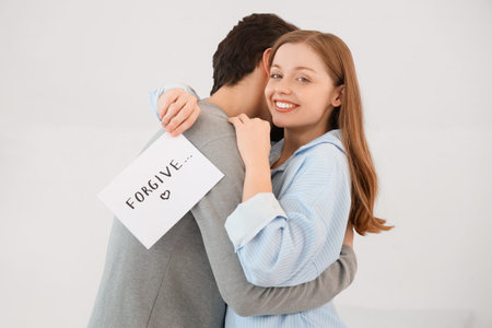 Young woman holding paper with the word FORGIVE and hugging her husband in bedroom