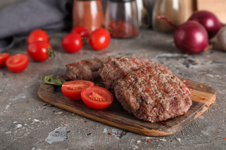 Wooden board with tasty meat cutlets, cut tomato and rosemary on gray grunge background