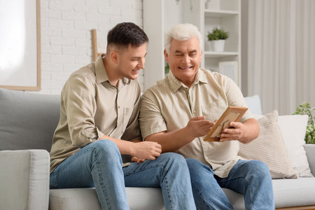 Young man and his father with photo frame sitting on sofa at homeの写真素材