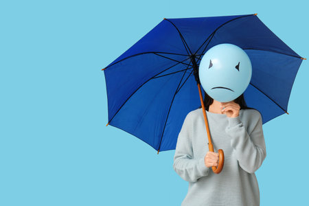 Young woman holding umbrella and balloon with sad face on blue background. Blue Monday conceptの写真素材