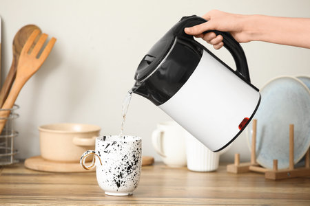 Woman pouring water from modern electric kettle into cup on counter in kitchen