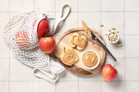 Wooden board with tasty apple slices, nut butter and peanuts on white tile backgroundの写真素材