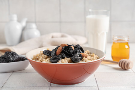 Bowl of tasty oatmeal with prunes, almonds and jar of honey on white tile backgroundの写真素材