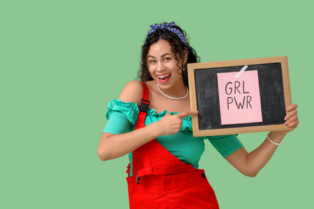 Young African-American pin-up woman pointing at GRL PWR sign on chalkboard against green background. Women's History Monthの写真素材