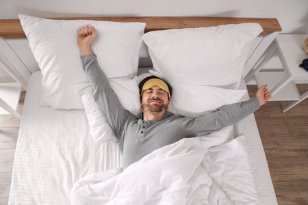 Handsome young man with sleep mask stretching in bed at homeの写真素材