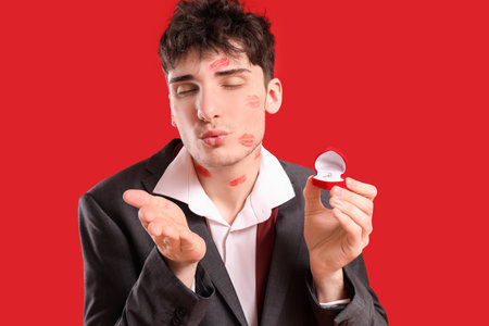 Handsome young man with engagement ring and lips marks on his face blowing kiss against red backgroundの写真素材