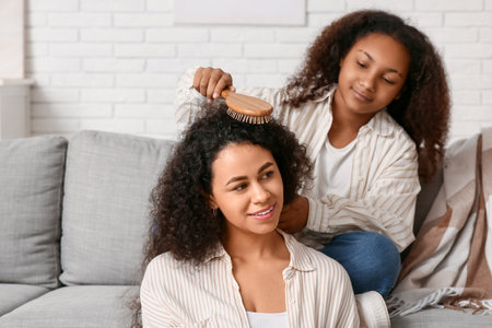 African-American girl brushing her happy mother's curly hair on sofa at homeの写真素材