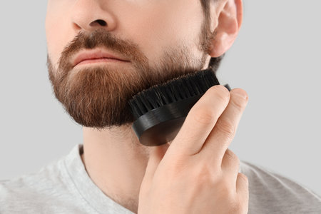 Handsome young man brushing his beard on gray background, closeupの写真素材