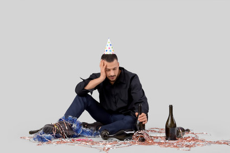 Young man with tinsel and bottles feeling unwell after party on light background