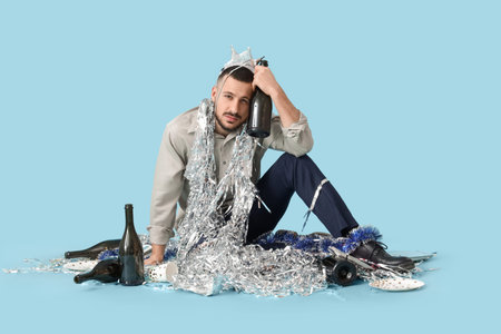 Young man with bottle and tinsel feeling unwell after party on blue background