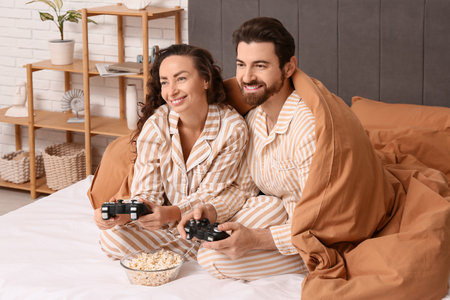 Happy couple in pajamas with blanket playing video game on bed at homeの写真素材