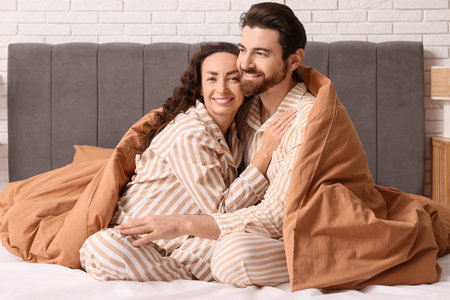 Happy couple in pajamas with blanket hugging on bed at homeの写真素材