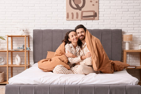 Happy couple in pajamas with blanket sitting on bed at homeの写真素材