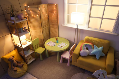 Interior of stylish children's room with cozy armchair, toys, glowing lights and folding screen at nightの写真素材
