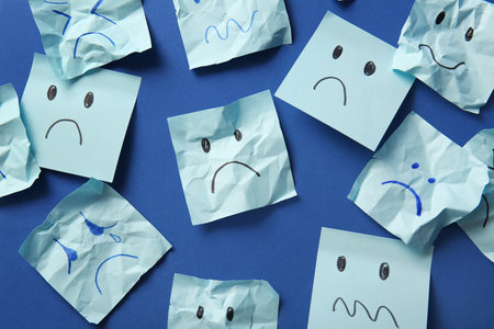 Paper sheets with drawn sad faces on blue background, top viewの写真素材