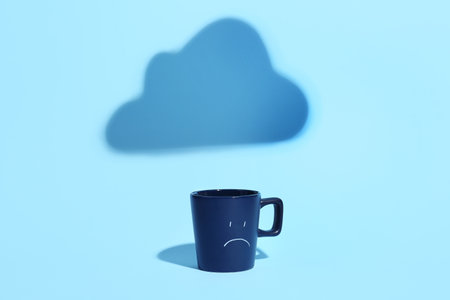 Cup with drawn sad face and shadow of cloud on blue backgroundの写真素材