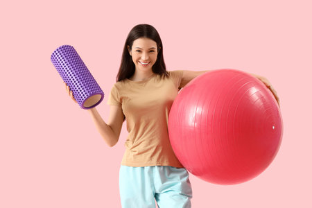 Young woman with foam roller and fitball on pink backgroundの写真素材
