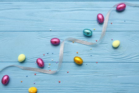 Ribbon with chocolate Easter eggs in foil and sprinkles on blue wooden background