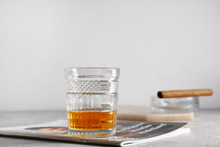 Glass of cold rum, cigar and magazine on table against gray background. Closeup