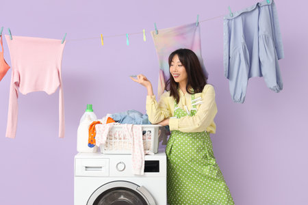 Happy young Asian woman with hanging laundry, basket and clothespins near washing machine on purple backgroundの写真素材