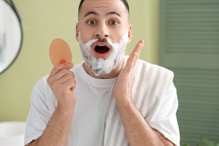 Shocked young man with soap washing his beard in bathroom, closeupの写真素材