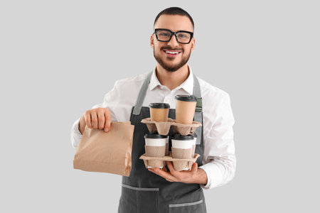 Young male barista with paper cups of coffee and food on gray backgroundの写真素材