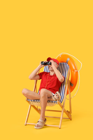 Male lifeguard with rescue ring looking through binoculars in deck chair on yellow backgroundの写真素材