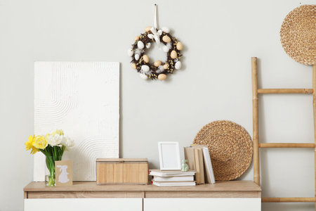 Chest of drawers with tulips, books and Easter wreath on light wall in room