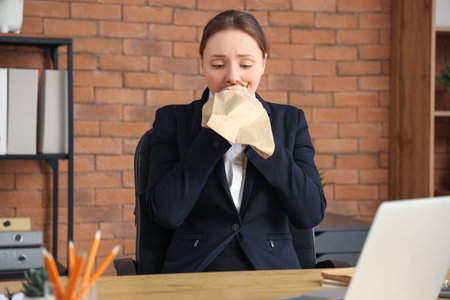 Young businesswoman with paper bag having panic attack at table in officeの写真素材
