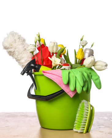 Bucket with bottles of detergents, cleaning tools and spring flowers on table against white backgroundの写真素材