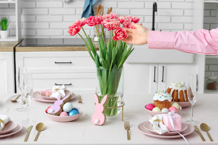 Female hands preparing beautiful Easter table setting with tulip flowers in kitchen. Closeup