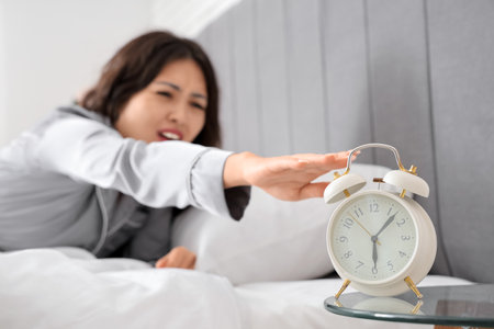 Displeased young woman turning off alarm clock on table in bedroom, closeupの写真素材