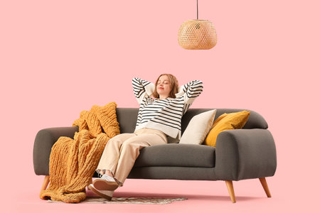 Beautiful young woman sitting on stylish gray sofa against pink backgroundの写真素材