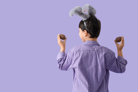 Cute boy in bunny ears with sweet chocolate eggs on lilac background, back view. Easter celebration