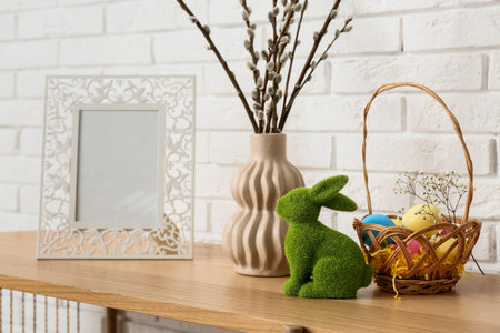 Easter rabbit with basket of colorful eggs and willow branches in vase on commode near white brick...