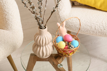 Willow branches in vase with wicker basket of colorful eggs and paper rabbit on table