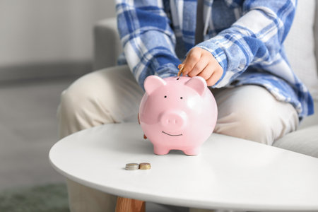 Young woman putting coin into piggy bank on table at home, closeupの写真素材