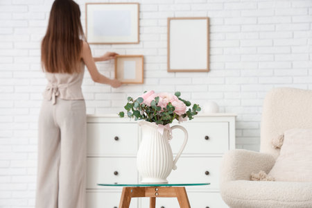 Young woman hanging frames on wall in stylish living room, focus on vase with bouquet of beautiful ranunculus flowersの写真素材