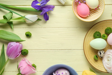 Frame made of different flowers and painted eggs for Easter celebration on yellow wooden background